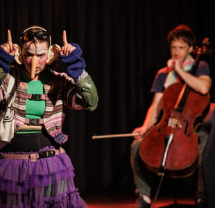 To utøvere på scenen: fargerikt kledd person som gestikulerer, og cellist som spiller et instrument i bakgrunnen. Teatralsk belysning.