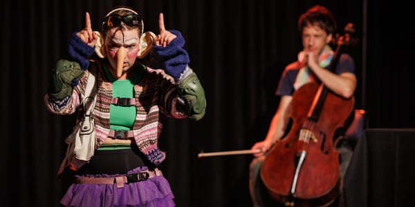 To utøvere på scenen: fargerikt kledd person som gestikulerer, og cellist som spiller et instrument i bakgrunnen. Teatralsk belysning.