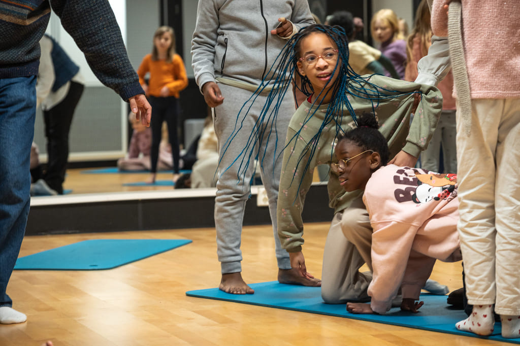 Flere barn står og krabber står på en blå matte i et aktivitetsrom. Det er speil på veggene.