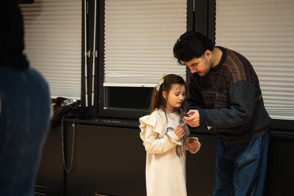 En voksen står ved siden av et barn i et rom med mørke vegger og gulv, mens barnet ser på mobilen han holder.