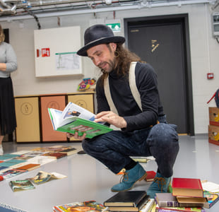 En person sitter på huk ved et bord fylt med bøker inne i et rom med grå vegger og en dør i bakgrunnen.