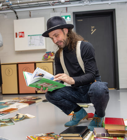 En person sitter på huk ved et bord fylt med bøker inne i et rom med grå vegger og en dør i bakgrunnen.