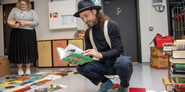 En person sitter på huk ved et bord fylt med bøker inne i et rom med grå vegger og en dør i bakgrunnen.