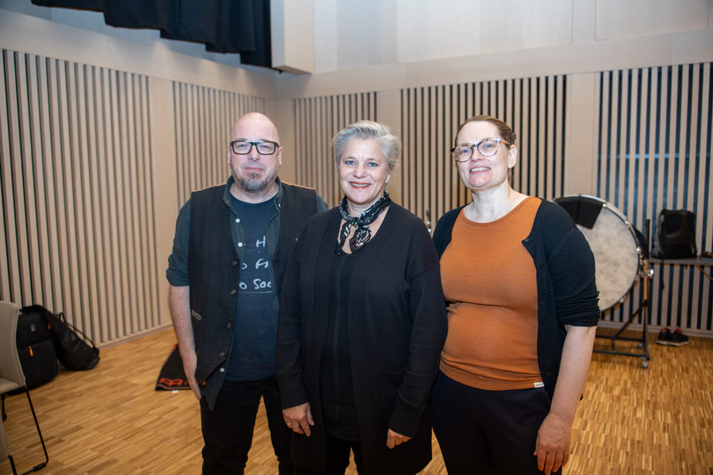 Tre personer står side om side i et rom med lyse trepaneler i bakgrunnen.