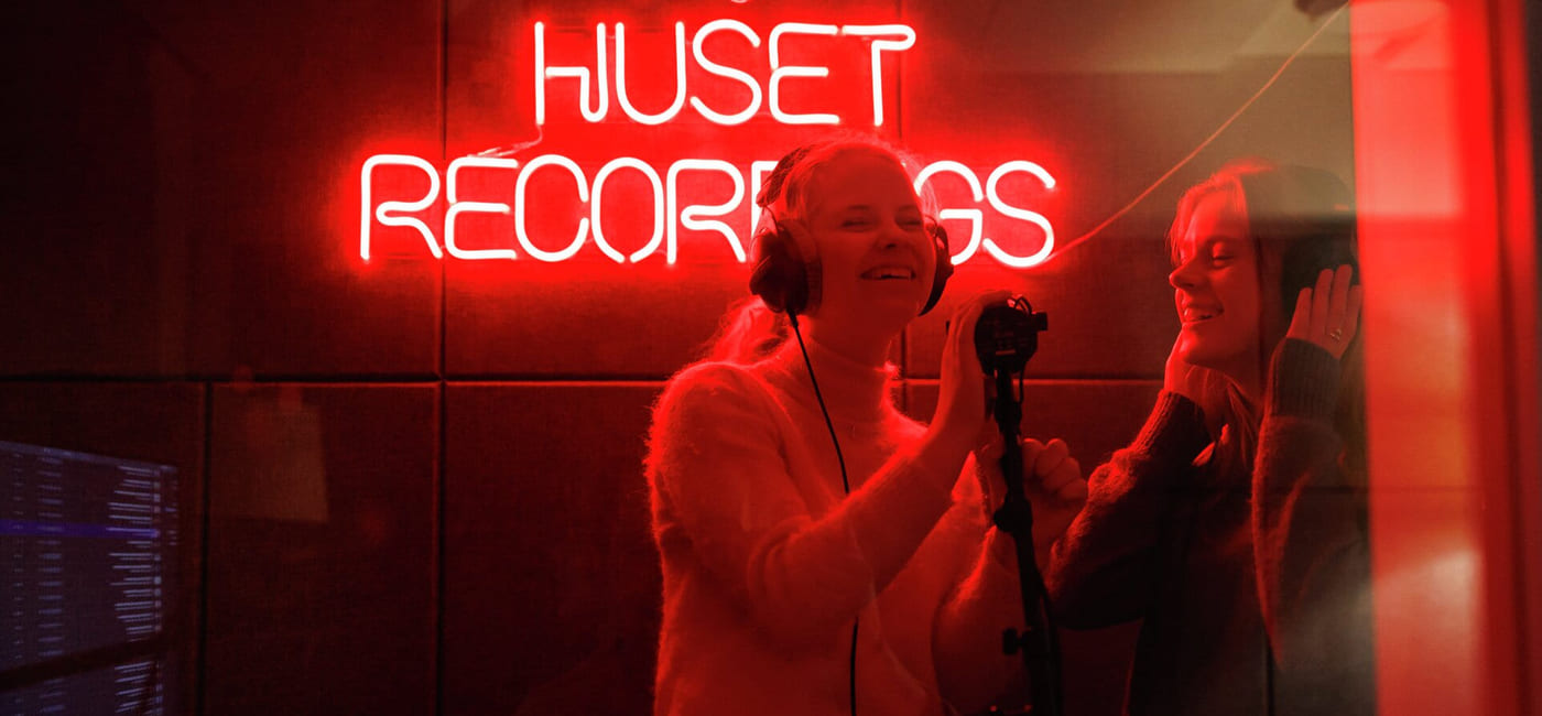 To personer i et innspillingsstudio foran et rødt neonskilt som sier «HUSET RECORDINGS».