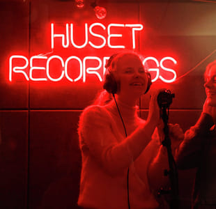 To personer i et innspillingsstudio foran et rødt neonskilt som sier «HUSET RECORDINGS».