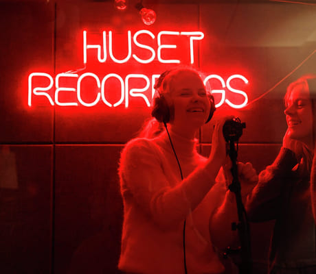 To personer i et innspillingsstudio foran et rødt neonskilt som sier «HUSET RECORDINGS».