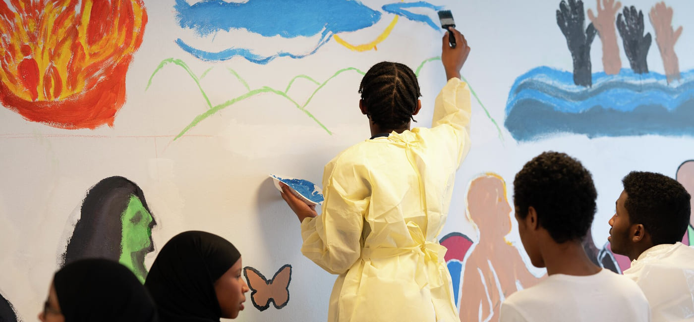 Person i gult maler et veggmaleri mens andre ser på. Fargerike bilder inkluderer ild, skyer, hender og figurer på veggen.