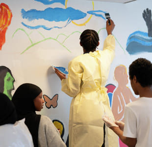 Person i gult maler et veggmaleri mens andre ser på. Fargerike bilder inkluderer ild, skyer, hender og figurer på veggen.