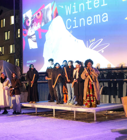 En gruppe mennesker står på en utendørsscene foran en stor skjerm med teksten «Winter Cinema».