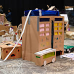 Pappkreasjoner, inkludert bygninger og kjøretøy spredt utover gulvet. Barnekunstprosjekt med esker, tape og fargerik dekorasjon i kreativ montering.