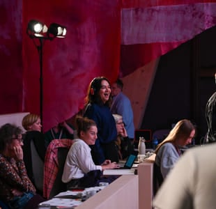 Folk som samhandler i et fargerikt produksjonsstudio eller arrangementslokale med rød bakgrunn og scenebelysning.