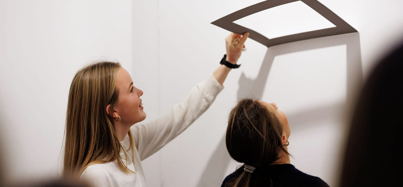 To unge kvinner står ved en vegg. Den ene holder opp en ramme. De ser begge opp mot den.