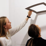 To unge kvinner står ved en vegg. Den ene holder opp en ramme. De ser begge opp mot den.