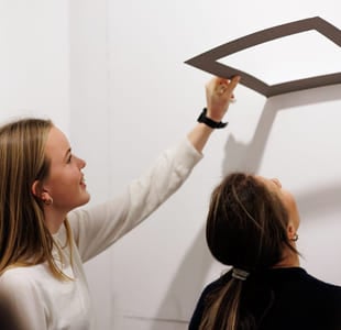 To unge kvinner står ved en vegg. Den ene holder opp en ramme. De ser begge opp mot den.
