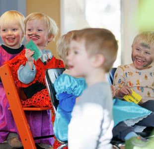 Barnehagebarn sitter på stoler og står. De ser på noe mens de ler. De har kledd seg ut.