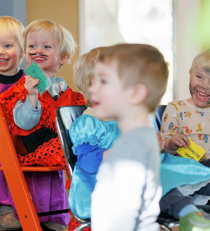 Barnehagebarn sitter på stoler og står. De ser på noe mens de ler. De har kledd seg ut.