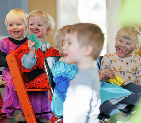 Barnehagebarn sitter på stoler og står. De ser på noe mens de ler. De har kledd seg ut.