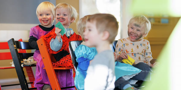 Barnehagebarn sitter på stoler og står. De ser på noe mens de ler. De har kledd seg ut.