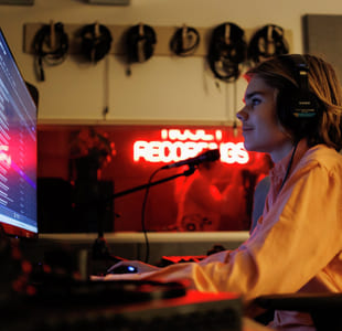 Person med hodetelefoner som jobber ved datamaskin i et svakt opplyst rom med neonskilt for «Recordings» og synlige lydkabler.