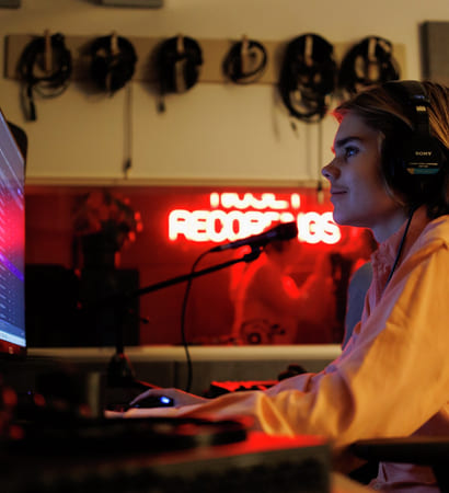 Person med hodetelefoner som jobber ved datamaskin i et svakt opplyst rom med neonskilt for «Recordings» og synlige lydkabler.