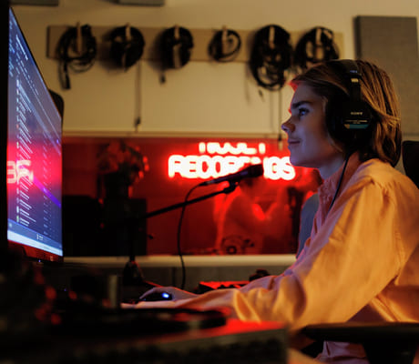 Person med hodetelefoner som jobber ved datamaskin i et svakt opplyst rom med neonskilt for «Recordings» og synlige lydkabler.