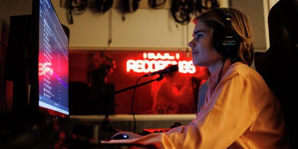 Person med hodetelefoner som jobber ved datamaskin i et svakt opplyst rom med neonskilt for «Recordings» og synlige lydkabler.