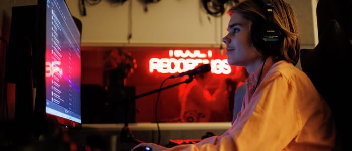 Person med hodetelefoner som jobber ved datamaskin i et svakt opplyst rom med neonskilt for «Recordings» og synlige lydkabler.