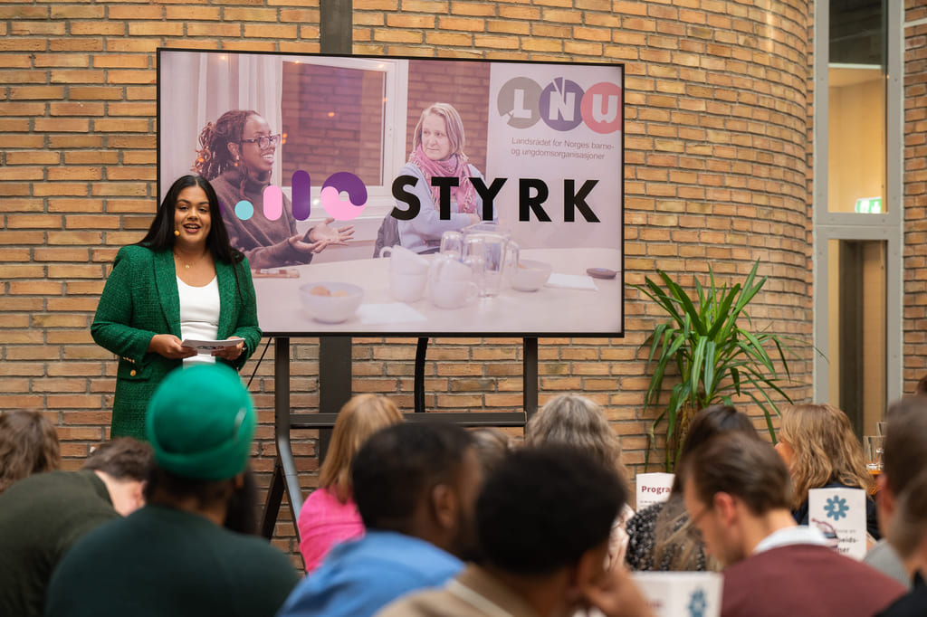 Presentasjon med foredragsholder foran skjerm og publikum som sitter og følger med.