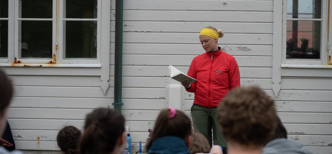 En kvinne i rød anorakk leser fra en bok for elever. De er ute.