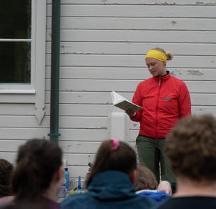 En kvinne i rød anorakk leser fra en bok for elever. De er ute.
