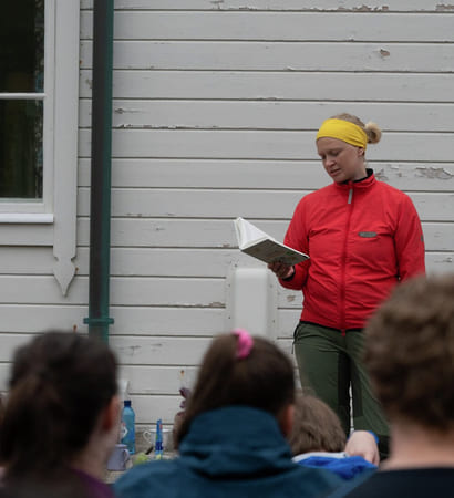 En kvinne i rød anorakk leser fra en bok for elever. De er ute.