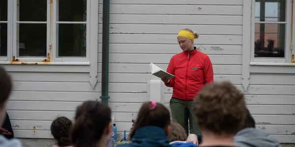 En kvinne i rød anorakk leser fra en bok for elever. De er ute.