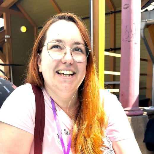 En kvinne i rosa t-skjorte blir intervjuet ved et grønt bord. Hun sitter i solskinnet.