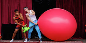 To utøvere på scenen med røde gardiner: én i stripete antrekk med lekepumpe, den andre blåser opp en gigantisk rød ballong.