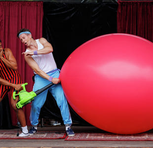To utøvere på scenen med røde gardiner: én i stripete antrekk med lekepumpe, den andre blåser opp en gigantisk rød ballong.