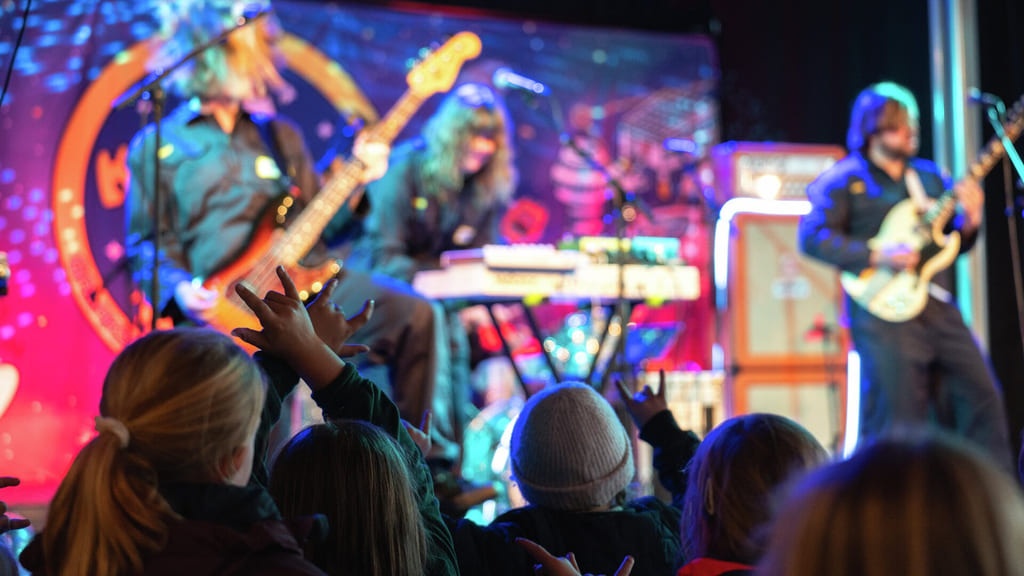 Rockeband opptrer på fargerik scene med gitarer og trommer. Entusiastisk publikum i forgrunnen rekker opp hendene og ser på showet.