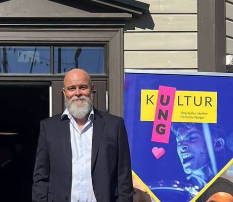 Smilende mann med skjegg står foran et hus med åpen dør. Ved siden står en rollup med teksten Ung kultur.