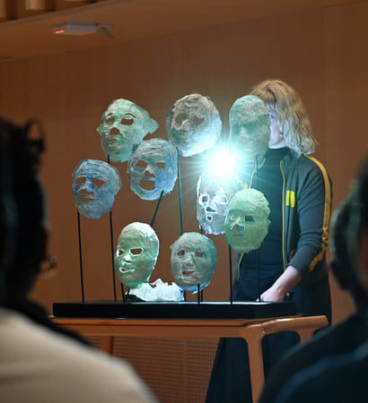 Kunstinstallasjon med opplyste, gjennomskinnelige masker på stativer, sett av tilskuere. En person i mørke klær står bak utstillingen.