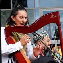 En musiker synger inn i en mikrofon mens han spiller på en rød Camac-harpe under en utendørsforestilling.