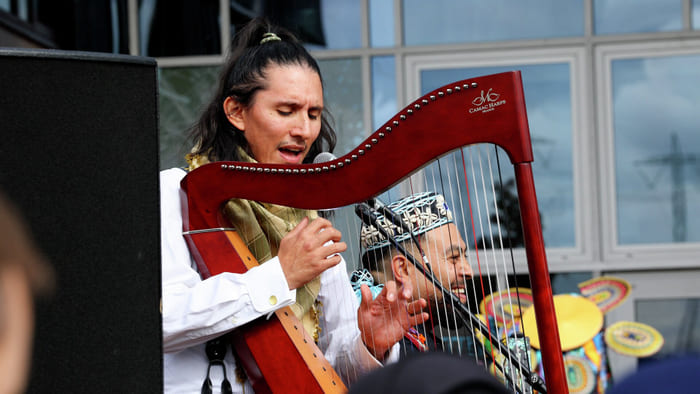 En musiker synger inn i en mikrofon mens han spiller på en rød Camac-harpe under en utendørsforestilling.