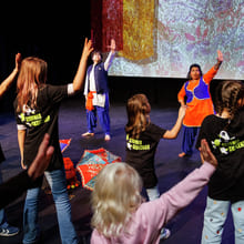Barn i Stovner Skolekor-skjorter rekker opp hendene under en kulturell danseforestilling med to tradisjonelt kledde instruktører på scenen.
