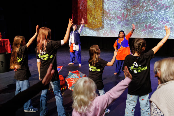 Barn i Stovner Skolekor-skjorter rekker opp hendene under en kulturell danseforestilling med to tradisjonelt kledde instruktører på scenen.
