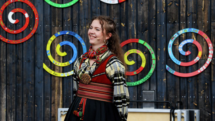 En smilende kvinne iført en fargerik tradisjonell nordisk folkedrakt står foran en trevegg med dekorative spiraler.