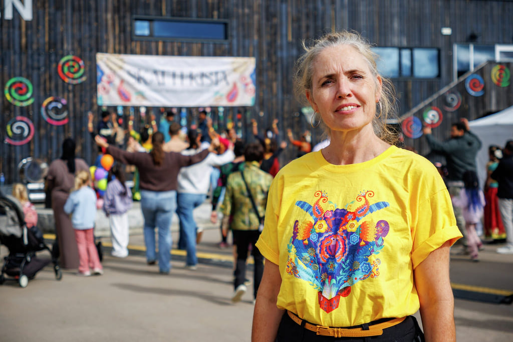 En smilende kvinne i en gul t-skjorte med grafikk står utendørs på et offentlig arrangement eller en parade med mange mennesker.