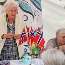 Eldre kvinner på et norskinspirert håndverksarrangement med norske flagg, flaggdekorasjoner og håndlagde gjenstander på bordet.