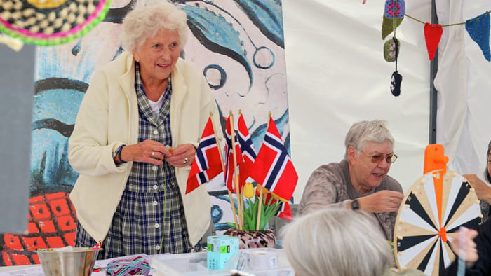 Eldre kvinner på et norskinspirert håndverksarrangement med norske flagg, flaggdekorasjoner og håndlagde gjenstander på bordet.