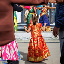 En ung jente i en rød og gullfarget indisk lehenga ser på tradisjonelle Bharatanatyam-dansere opptre på en kulturfestival.