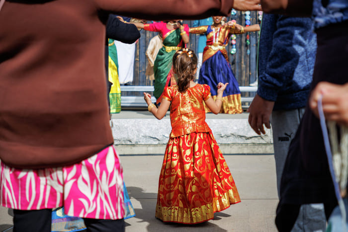 En ung jente i en rød og gullfarget indisk lehenga ser på tradisjonelle Bharatanatyam-dansere opptre på en kulturfestival.