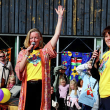 To kvinner i gule T-skjorter holder mikrofoner og vinker på en utendørsfestival med barn og fargerike ballonger.